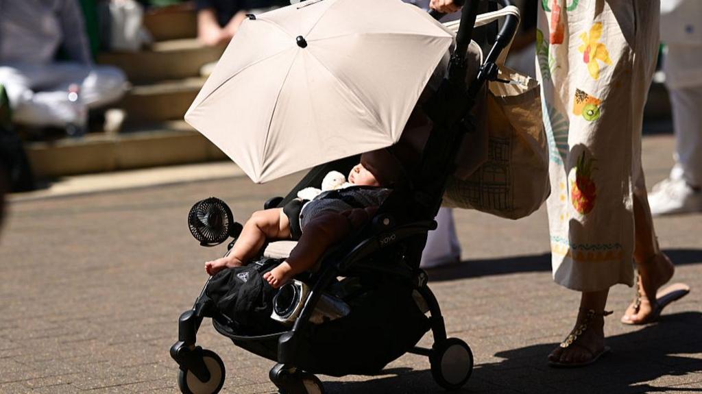  A baby is pushed in a pram with a fan and umbrella on day eleven of The Championships Wimbledon 2025 