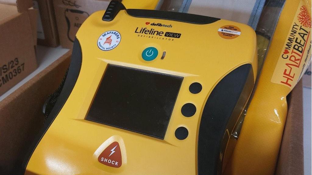 A yellow defibrillator with a screen in the middle with three black buttons on the right. At the top it has the writing 'Lifeline' with stickers around it. At the bottom, under the screen, is a red triangle with the words 'SHOCK'.