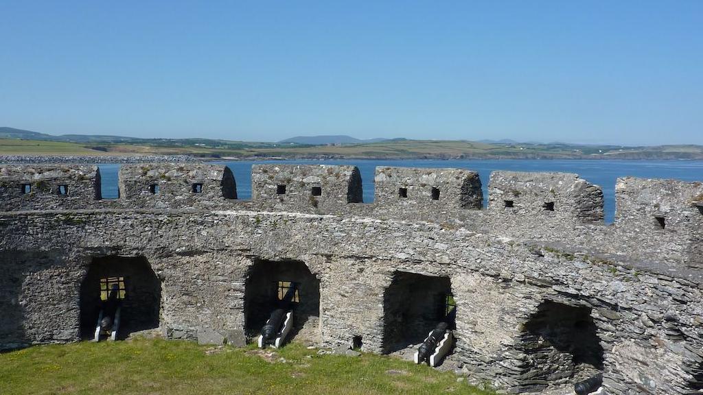 Fort Island from the inside, a stone fort wall arches around, with cannons facing out, there is grass inside, and behind you can see the blue sea and green land of the Isle of Man.
