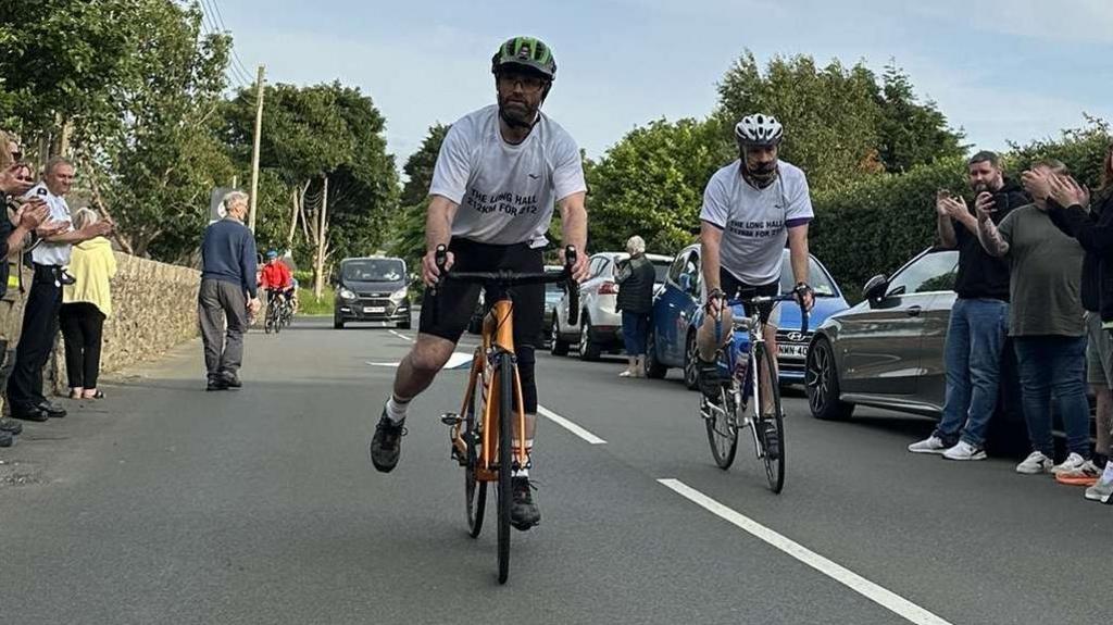 Two men wearing white t-shirts on bikes as they get clapped on at the finishing line on a road in Ballaugh.