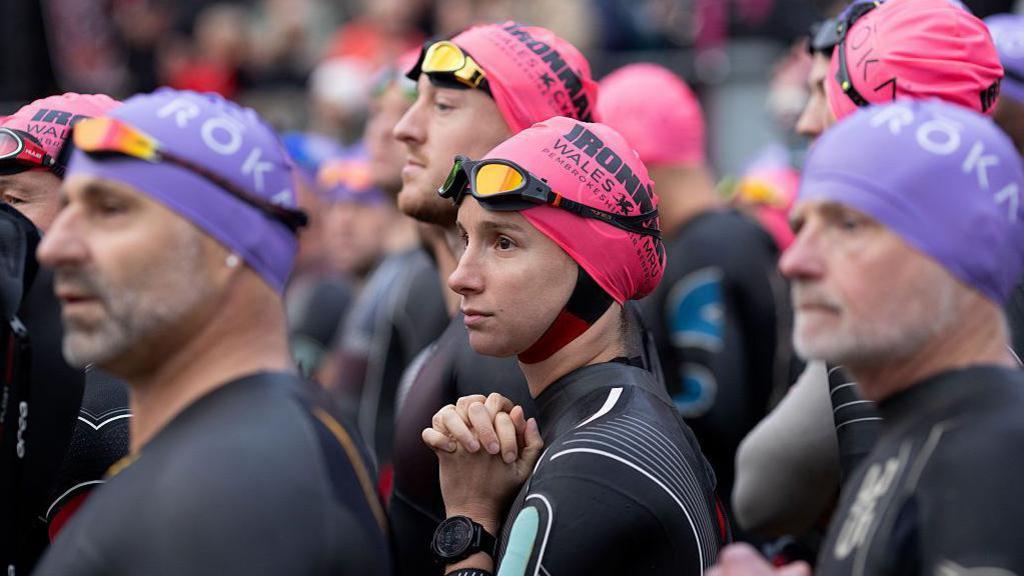 Men and women stand together wearing swimming caps, goggles and wetsuits