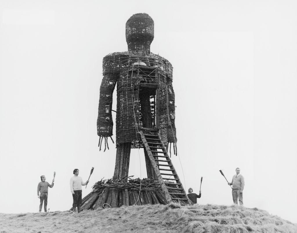 The Wicker Man. A large wicker man standing in a field with four people standing beside it with torches, waiting to set it alight.