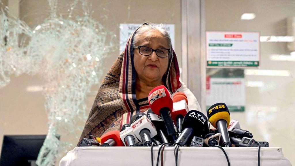 Sheikh Hasina stands in front of a bunch of microphones. Behind her is a shattered window.