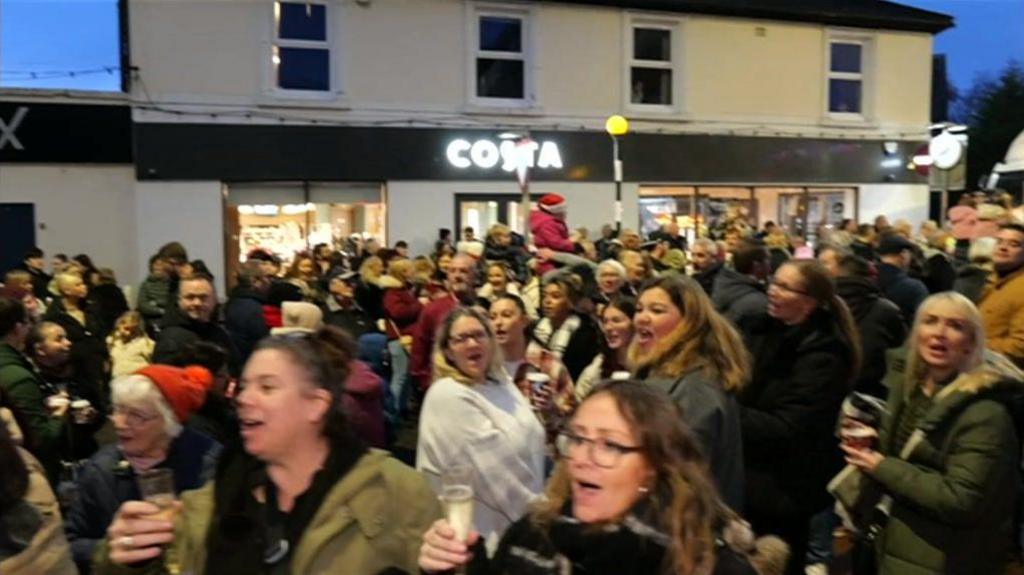 a crowd of people, dressed in winter coats, some wearing Christmas hats, some raising champagne flutes, stand outside singing Happy Birthday to Gladys Banks. A white building with a lit Costa sign is in the background and the sky is dark