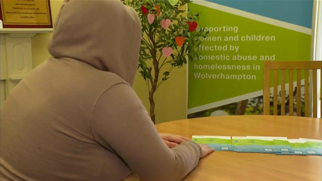 A woman wearing a light-brown hooded jacket is sat at a brown wooden table with a tree in the corner of the room next to a green, turquoise and white sign.