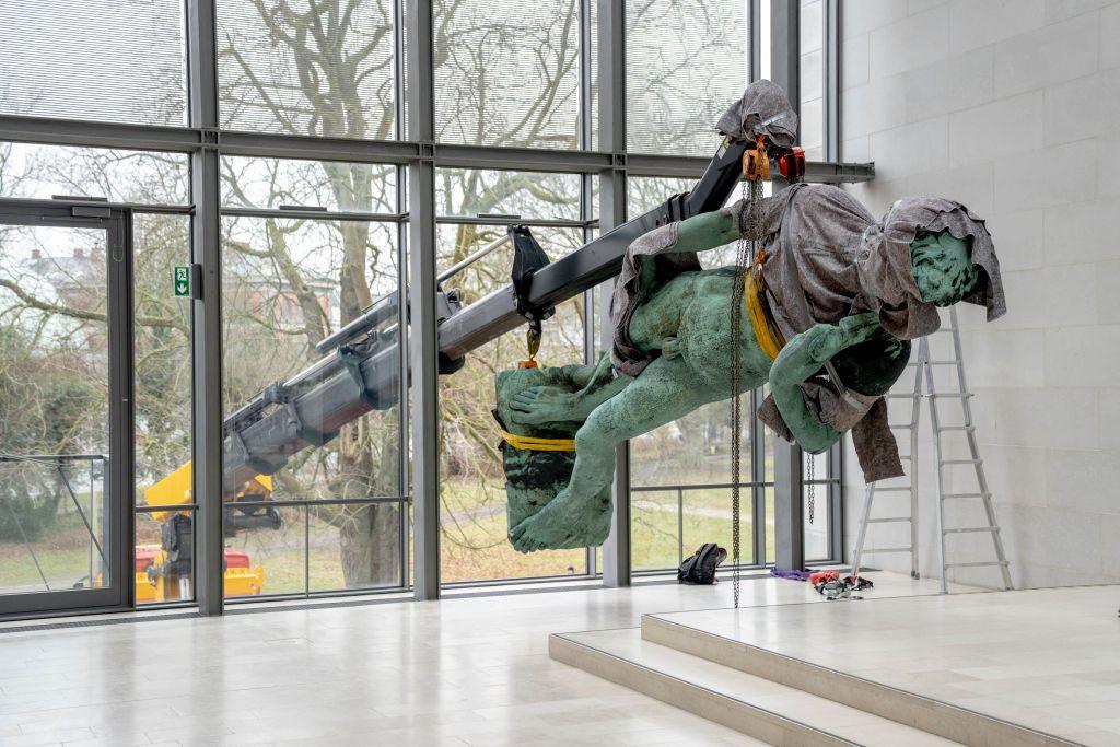 The statue David is lifted by crane through a large window of the National Gallery of Denmark. The statue is on its side, partially covered in a grey drape, as it is moved through the window