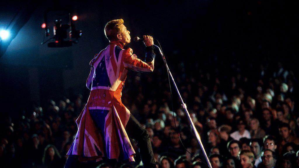 David Bowie performs in front of a large audience while wearing a long coat fashioned after the Union flag