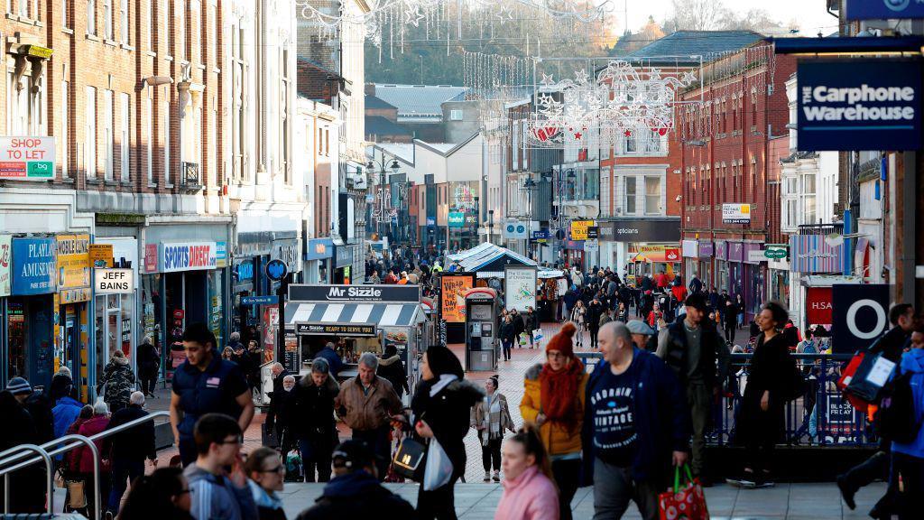 A busy Walsall town centre. Carphone Warehouse can be seen. There are lots of people walking around. It is Christmas time and there are lights. Most people have warm clothes on.