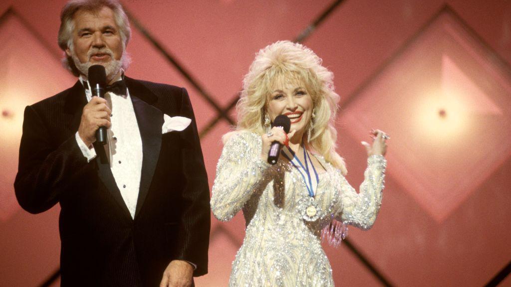 Kenny Rogers, wearing a black and white tuxedo, and Dolly Parton, wearing a bejewelled white and cream long sleeved dress, performing on stage with a pink background.