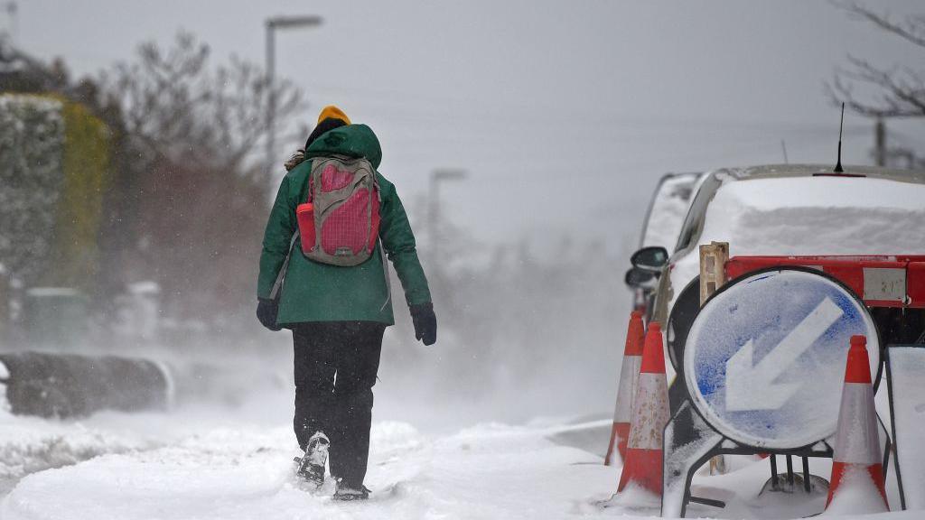 Person wearing thick coat and hat walking along a pavement covered in snow. 