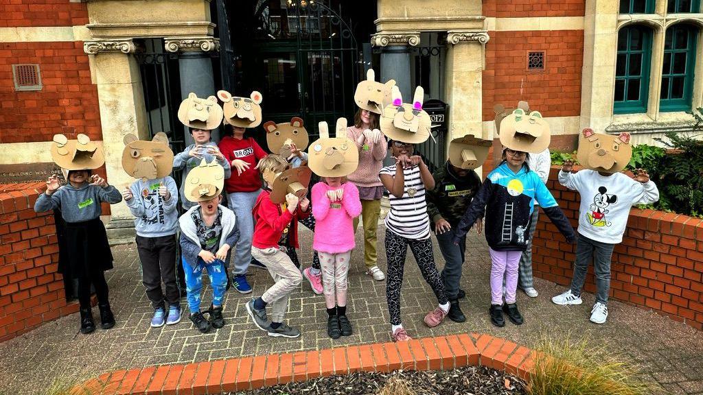 A group of children stand outside the Applecart Arts venue wearing handmade animal masks. They pose playfully in front of the historic building’s arched entrance.