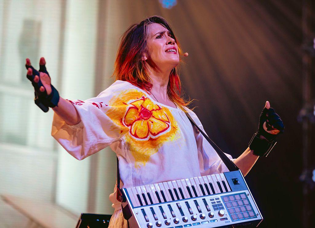 Imogen Heap performs on stage in Los Angeles earlier this year. She has her arms outstretched and is wearing a white top with an orange flower on it,, and black gloves.