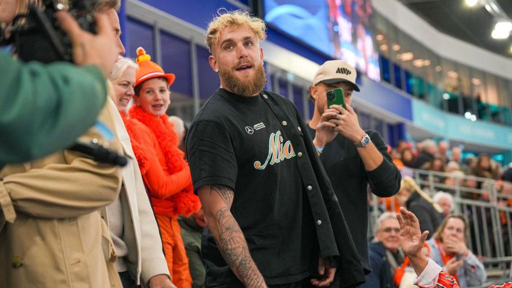 Jake Paul wearing a black tshirt and standing in a sports stadium as fans wave and take photos with their phones.