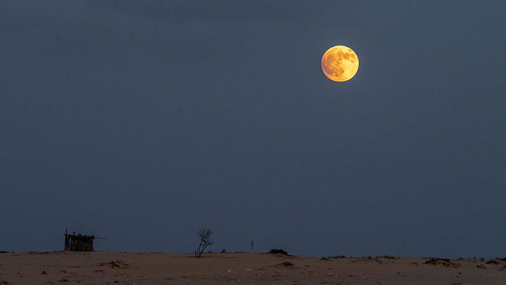 A bright red Moon high in a hazy, pale sky above a sandy desert