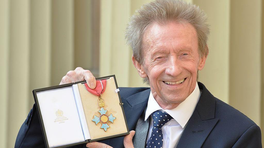 Denis Law holds up his CBE medal for the camera and smiles. He wears a dark suit and a spotted tie.