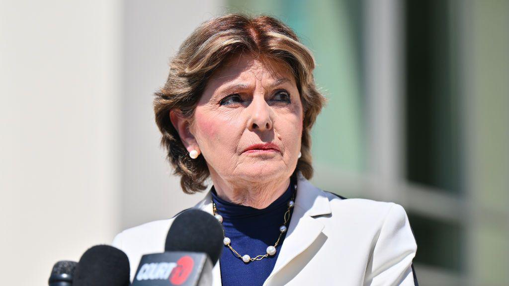 Gloria Allred. She has short brown hair, wearing pearl earrings and necklace, blue top and white blazer and standing in front of microphones.