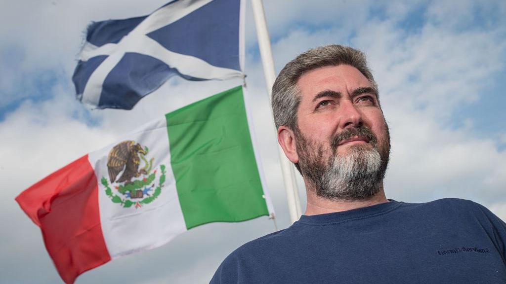 David Milne, a bearded man in a dark blue T-shirt stands in front of a Saltire and a Mexican flag.
