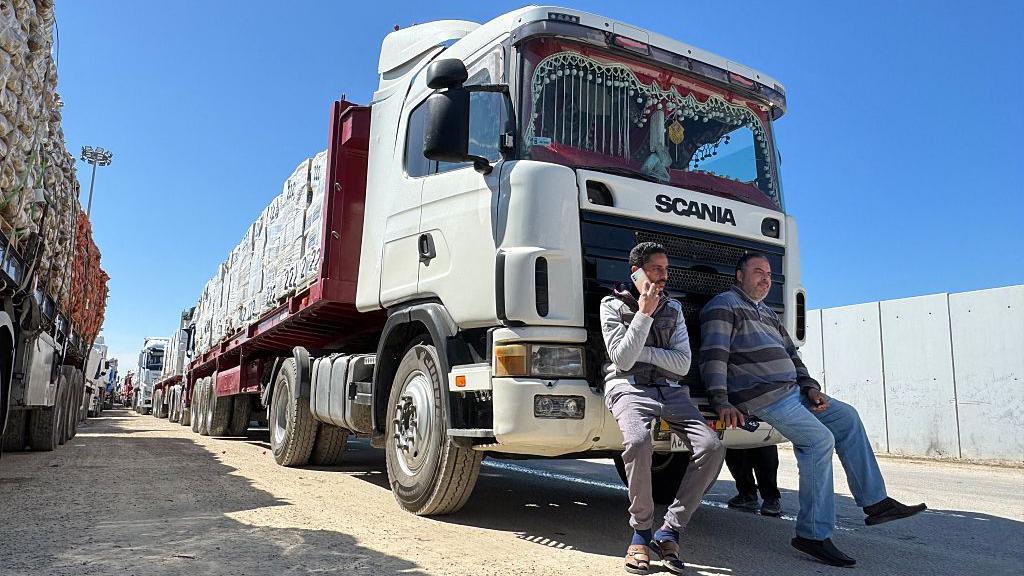 Two men sit on the front of a large lorry, looking a bit bored. The lorry is packed with aid parcels, and there is a long line of lorries behind it.