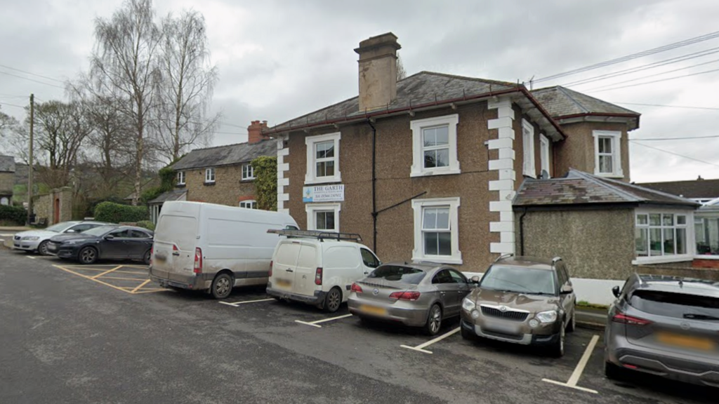 A two storey building with a brown cobbled exterior and white window pains. There are cars parked in the car park alongside it.