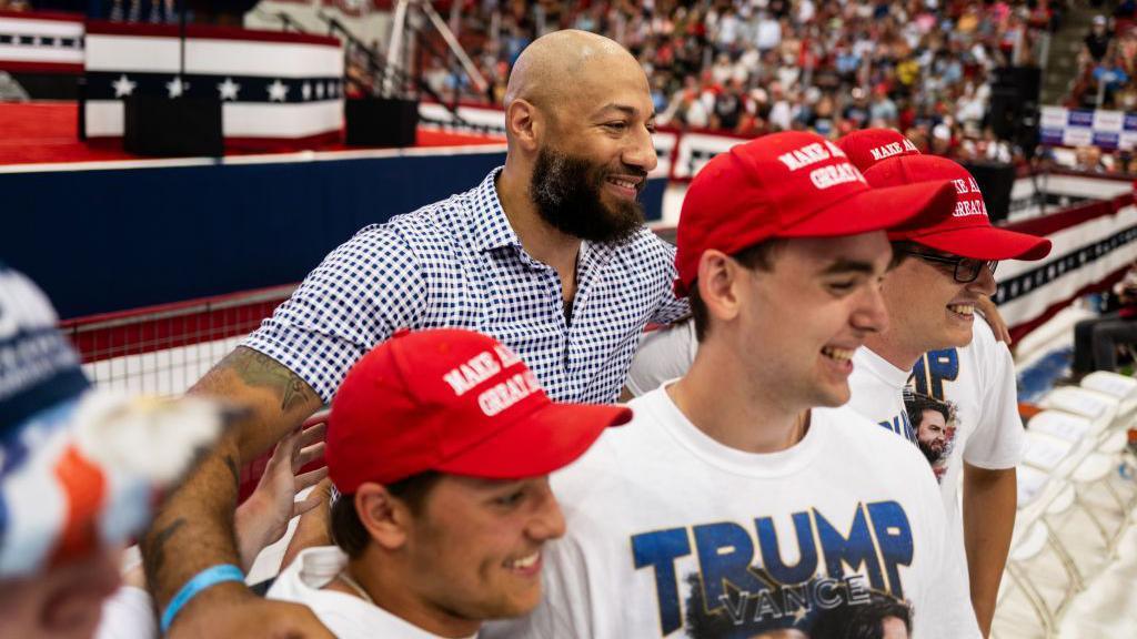 White in a blue checked shirt behind a line of supporters wearing red MAGA hats and Trump-Vance T-shirts