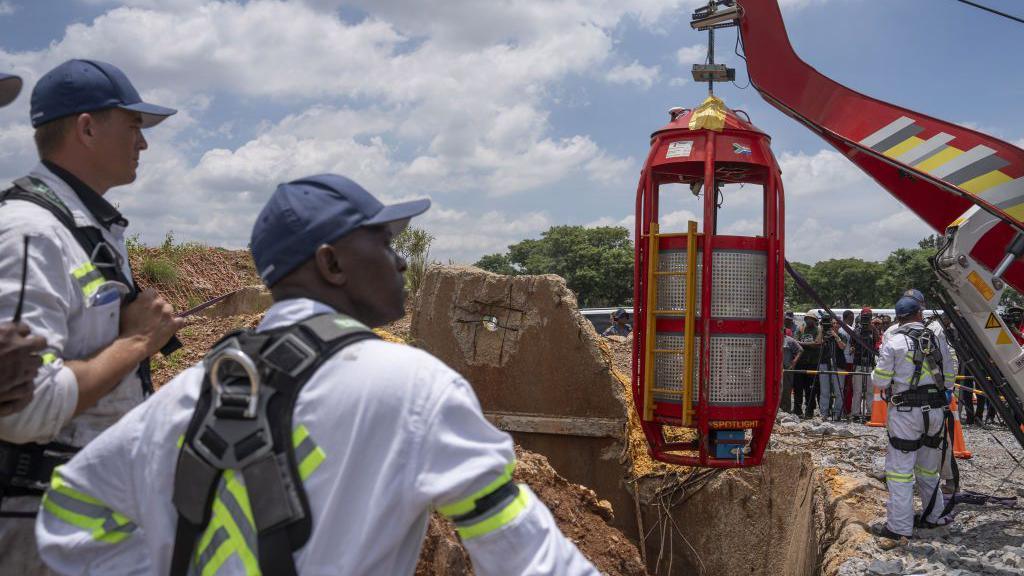 A special cage for hoisting people up from a mine shaft is readied at the top before being sent down. Workers in white overalls are looking on.