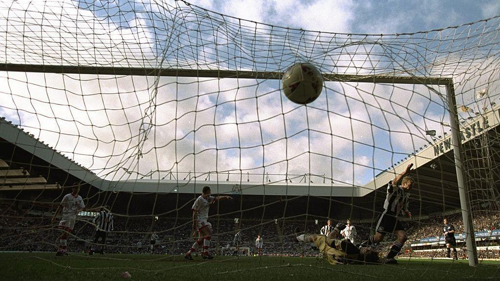 Pictured from behind the netting, Alan Shearer raises his right arm in celebration as he equalises for Newcastle United against Sunderland. Sunderland's goalkeeper is stricken on the ground as other players look on. The ground is filled with thousands of supporters.