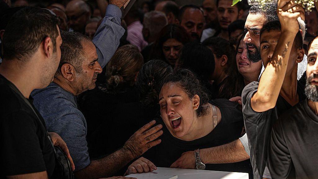 A woman cries leaning on top of a coffin as she is comforted by a man nearby as people standing behind her cry too