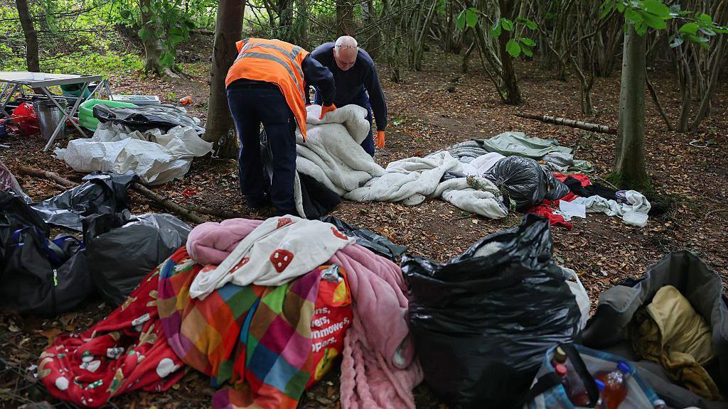 Two men piling duvets and blankets into black bin bags. There are blankets, duvets and bin bags strewn around the forest floor.