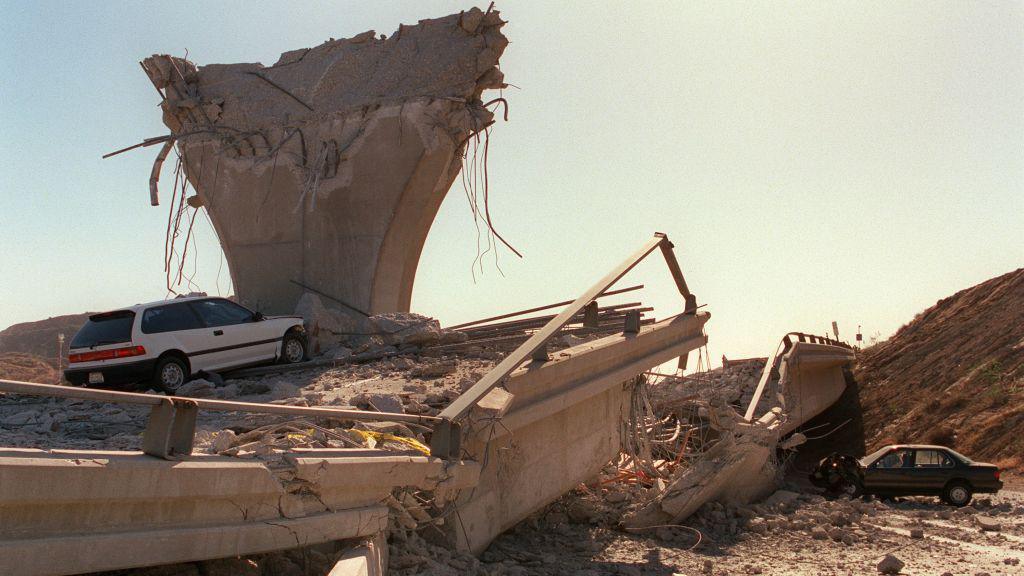 A highway has been turned into rubble after an earthquake, with an overpass split in half and two cars abandoned in the rubble