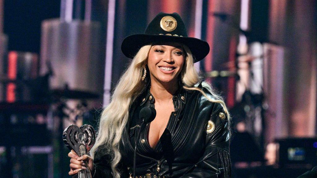 Beyoncé pictured on stage wearing a black cowboy hat, black jacket and holding a trophy. She has blonde hair and is smiling.