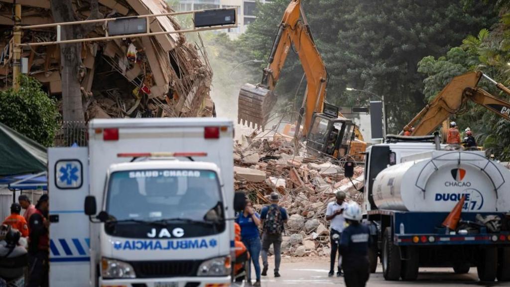 Trucks in a street in front of a large pile of debris which a yellow digger is on top of removing rubble from the pile 