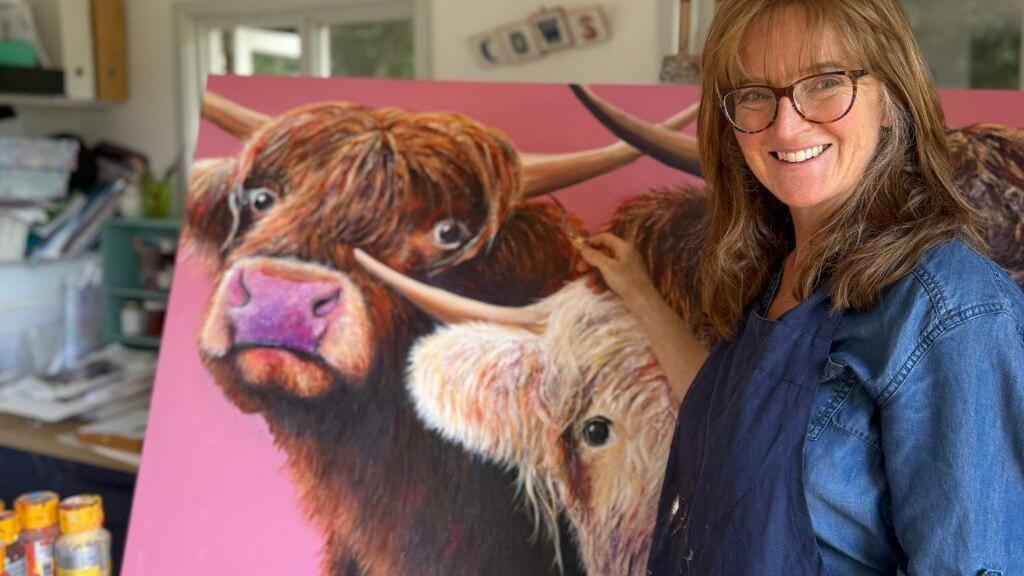 Jackie Spurrier is standing next to a canvas with highland cows painted on it in front of a pin background. Jackie is wearing large, brown-rimmed glasses, a blue jean jacket and has long brown hair. She is smiling at the camera.