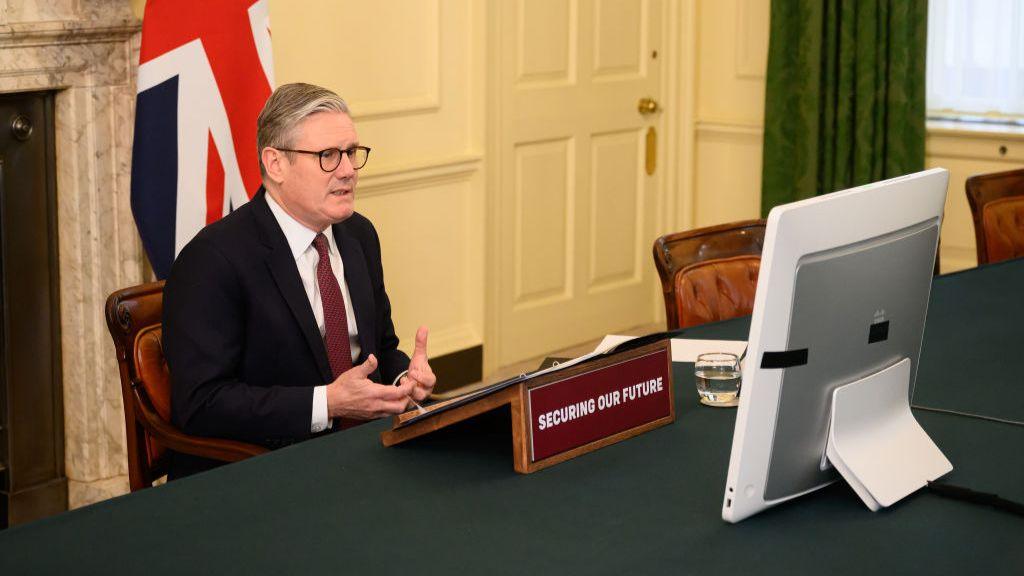 Starmer sits at his desk in a suit, speaking to a computer. 