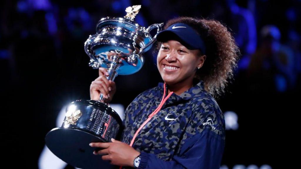 Naomi Osaka holds the Australian Open trophy in 2021. She is wearing a blue and grey tracksuit jacket with a pink zip, has a blue visor on her head and is smiling broadly.