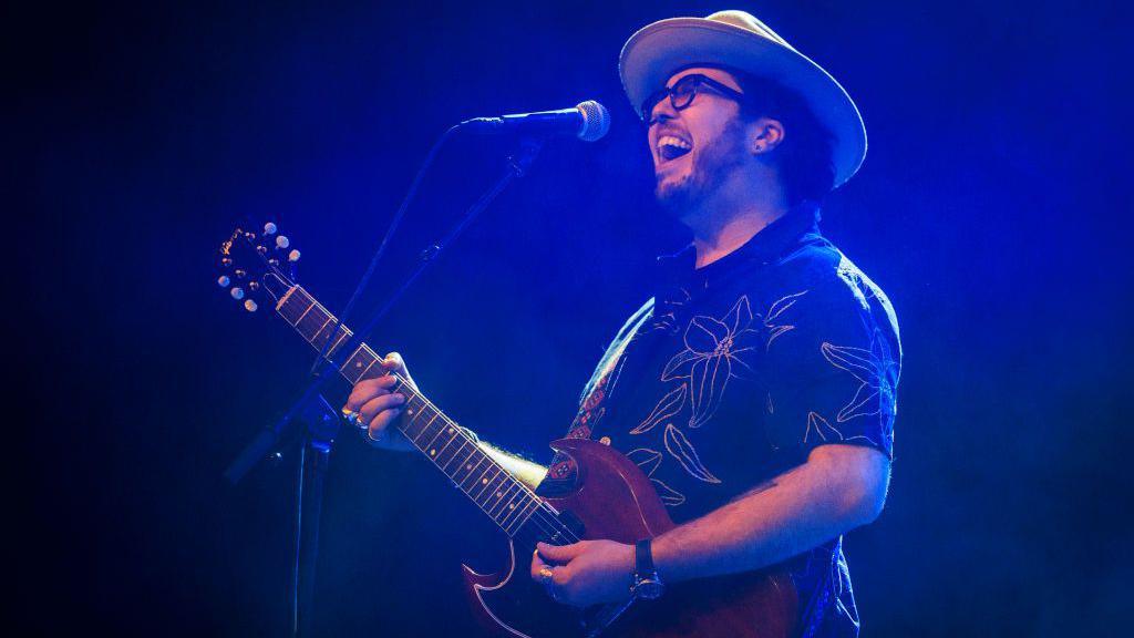 A singer with the band the Cinelli Brothers in sunglasses and a hat plays his guitar in a patterned shirt in front of a microphone