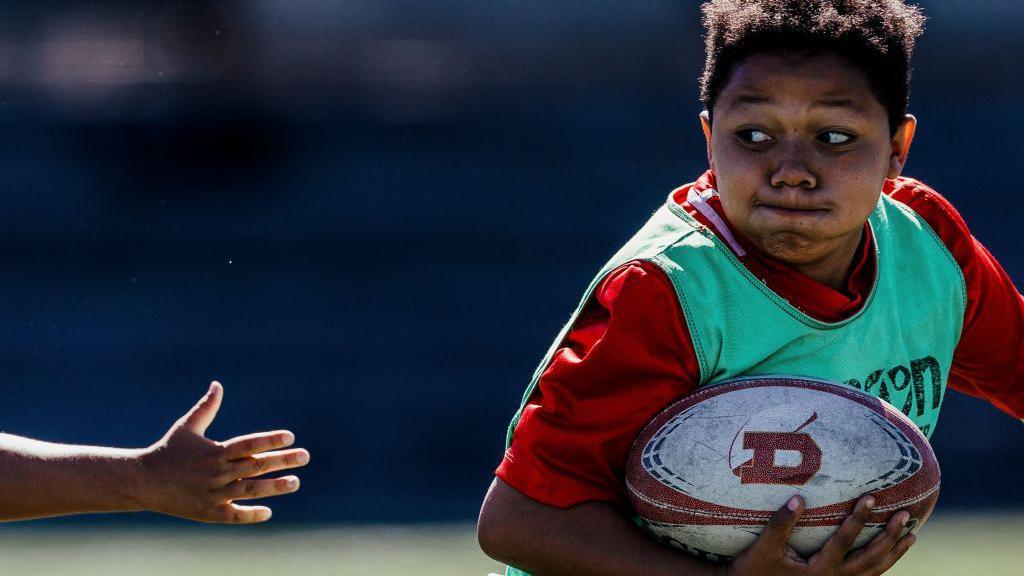 A young boy runs with a rugby ball. He looks to his right to see an opponent's hand trying to grab him. 