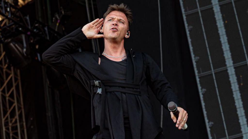 A man stands on a stage with one hand to his ear, a microphone held at his side in the other. He's wearing a black robe-style garment that's fastened with the type of clips usually found on backpacks. 