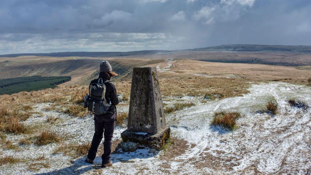 A person standing next to a trig point on a bare moor lightly dusted in snow