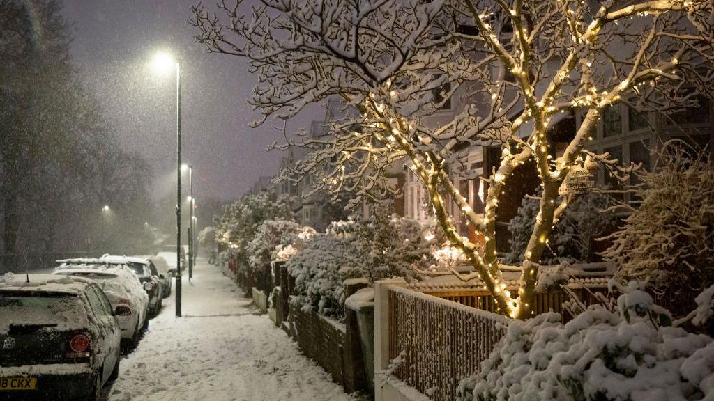 A street scene at night with lampposts lighting up falling snow with a blanket of snow on the ground, cars and trees.  Some Christmas lights can be seen in one of the trees