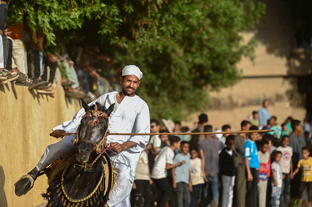 A man brandishes a wooden baton on horseback. A crowd behind him watches.