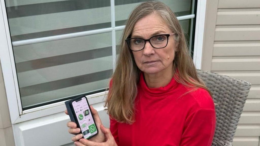 Felicity Campbell wears a red long-sleeved top and sits in a grey wicker chair in front of a window. She is holding up a mobile phone and looking into the camera