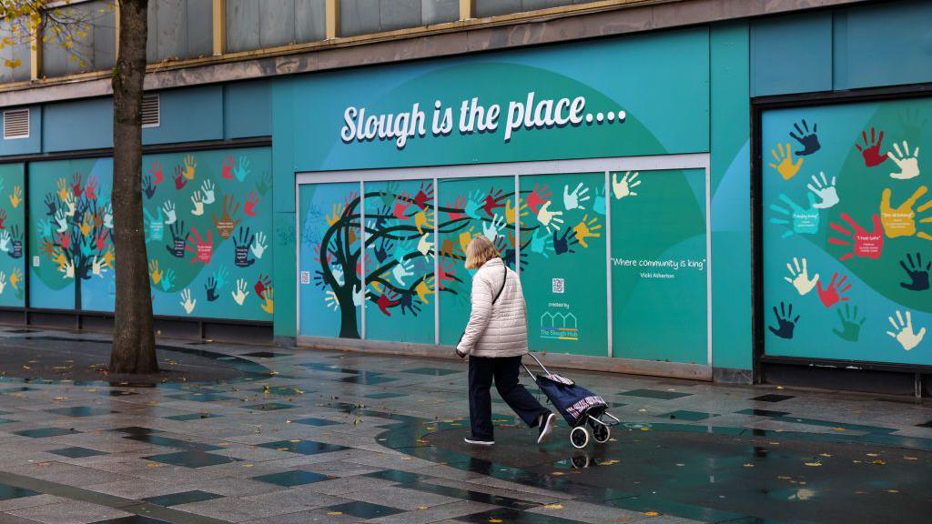 A woman with short, blonde hair with a shopping trolley which she is pulled walks on a wet, paved area in front of a boarded up shop which says "Slough is the place" and includes pictures of handprints in various colours and a painted, windswept tree, which also has various colours of handprints on it where branches would normally be 