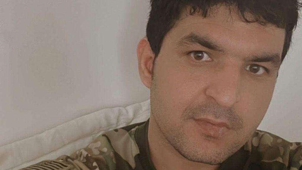 Rahmanullah Lakanwal in a selfie against a white background. He is wearing a military shirt in green and brown camouflge print