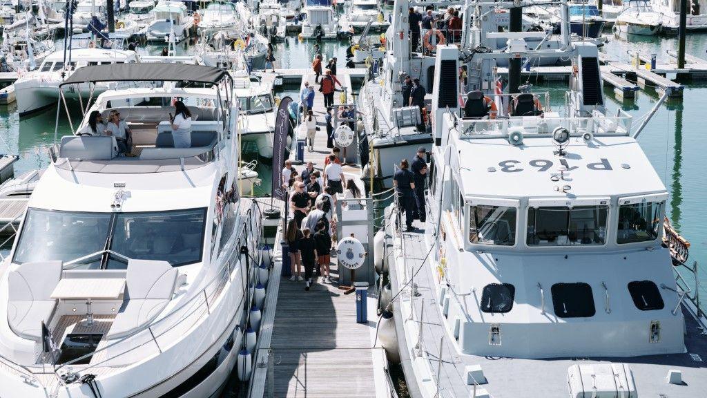 High angle shot of a marina pier in the sunshine. On either side of the pier are two superyachts. People can be seen stood on the pier looking at the boats