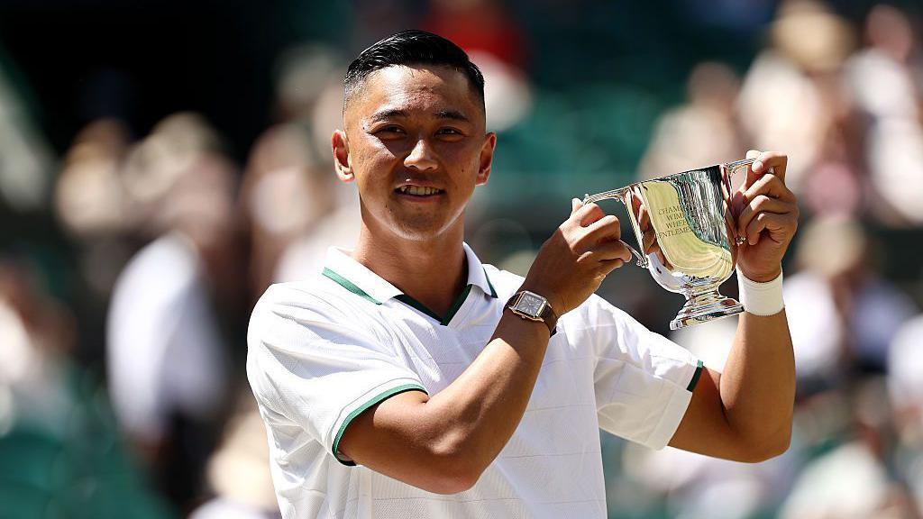Tokito Oda of Japan poses for a photo with his trophy. He has black hair and is wearing a white top with green details on the collar and sleeves. He is wearing a watch with a brown strap and a white sweatband on the other arm. He is holding up a silver trophy.