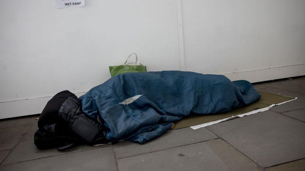 An unidentifiable person huddled in a sleeping bag on a pavement, with a rolled up coat over their face.