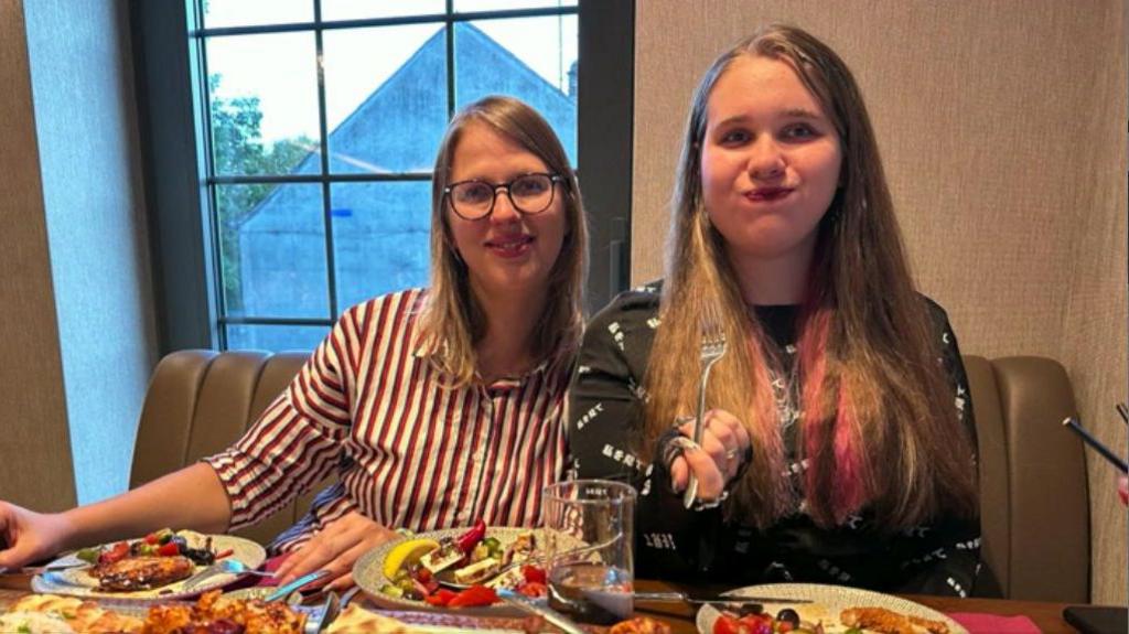 Two women sit at a table with plates of food in front of them. The woman on the right holds a fork and has puffed out her cheeks.