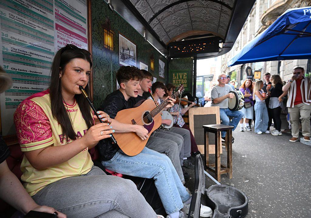 A woman in a yellow sports top and jeans playing a tin whistle with other musicians behind her at a bus stop. 