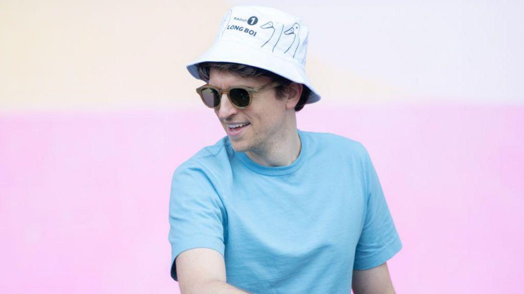 Greg James performs a DJ set in front of a pastel pink backdrop on-stage at Big Weekend. He wears a light blue t-shirt, sunglasses, and a white bucket hat printed with line-drawn pictures of a duck, the Radio 1 logo and the words "Long Boi" in black letters.
