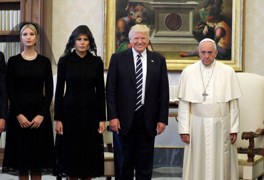 Ivanka and Melania, wearing dark clothes and black veils, stand next to Donald Trump, wearing a black suit and striped tie and beaming at the camera. Next to them is Pope Francis, dressed in white papal robes, with a sombre expression. 